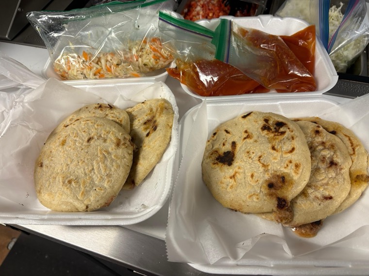 Two boxes of pupusas on the counter with curtido and red salsa alongside